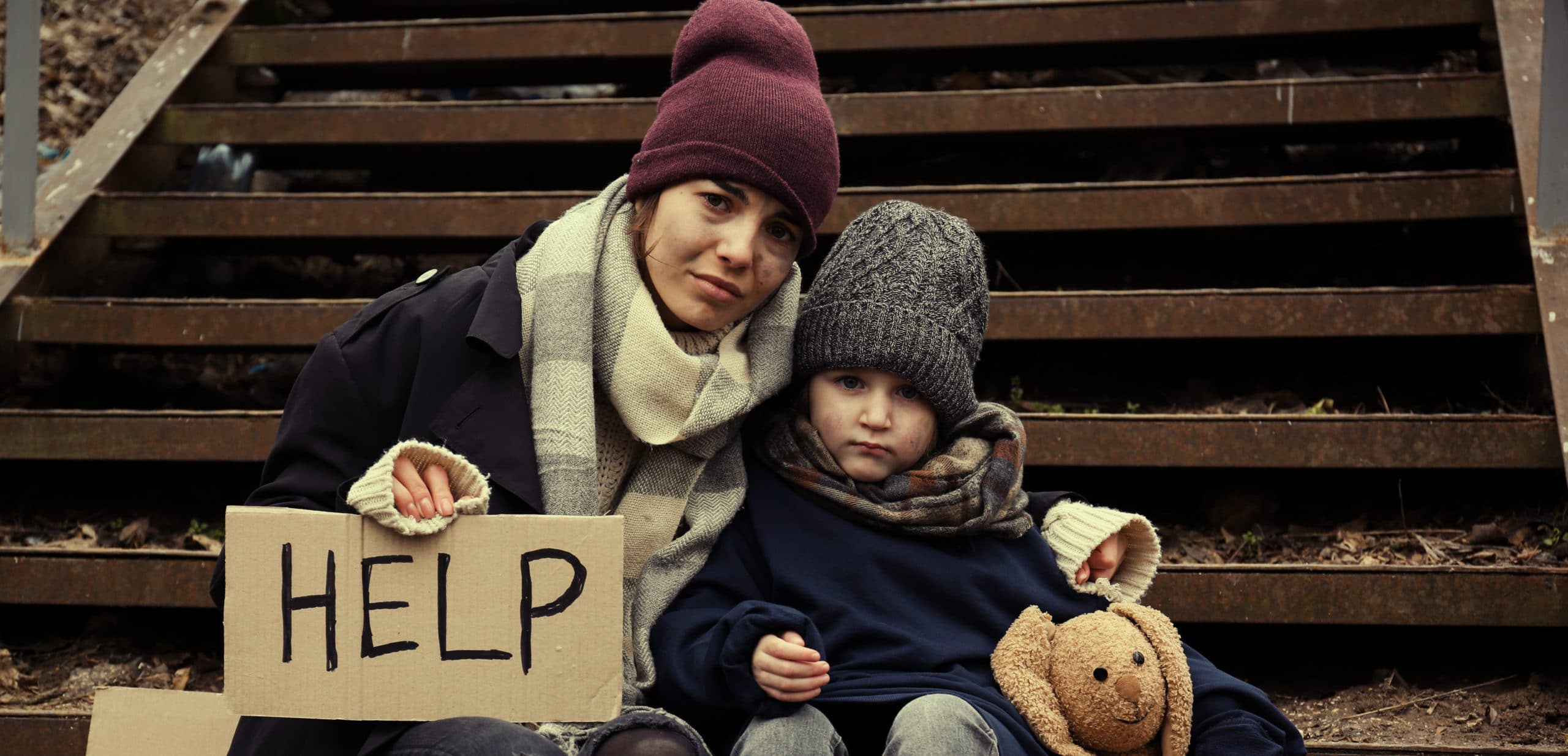 Poor mother and daughter with HELP sign sitting on stairs outdoors
