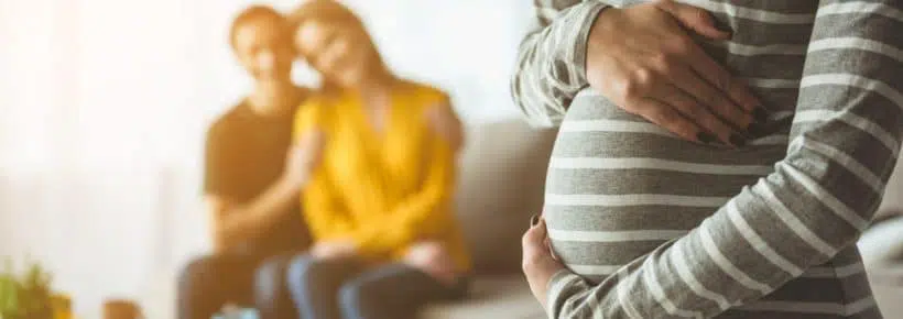 Focus on pregnant woman touching her belly. Happy married couple are sitting on sofa and embracing on background. Surrogacy concept