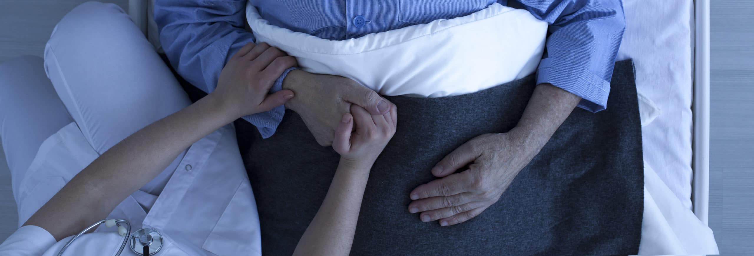 Photo from the top of nurse holding older sick man's hand who lies in hospital bed