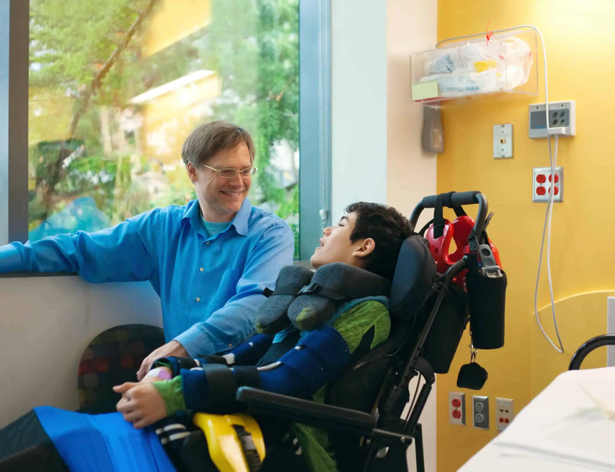 why euthanasia is wrong - Smiling father sitting next to disabled son in wheelchair by hospital bed, talking together