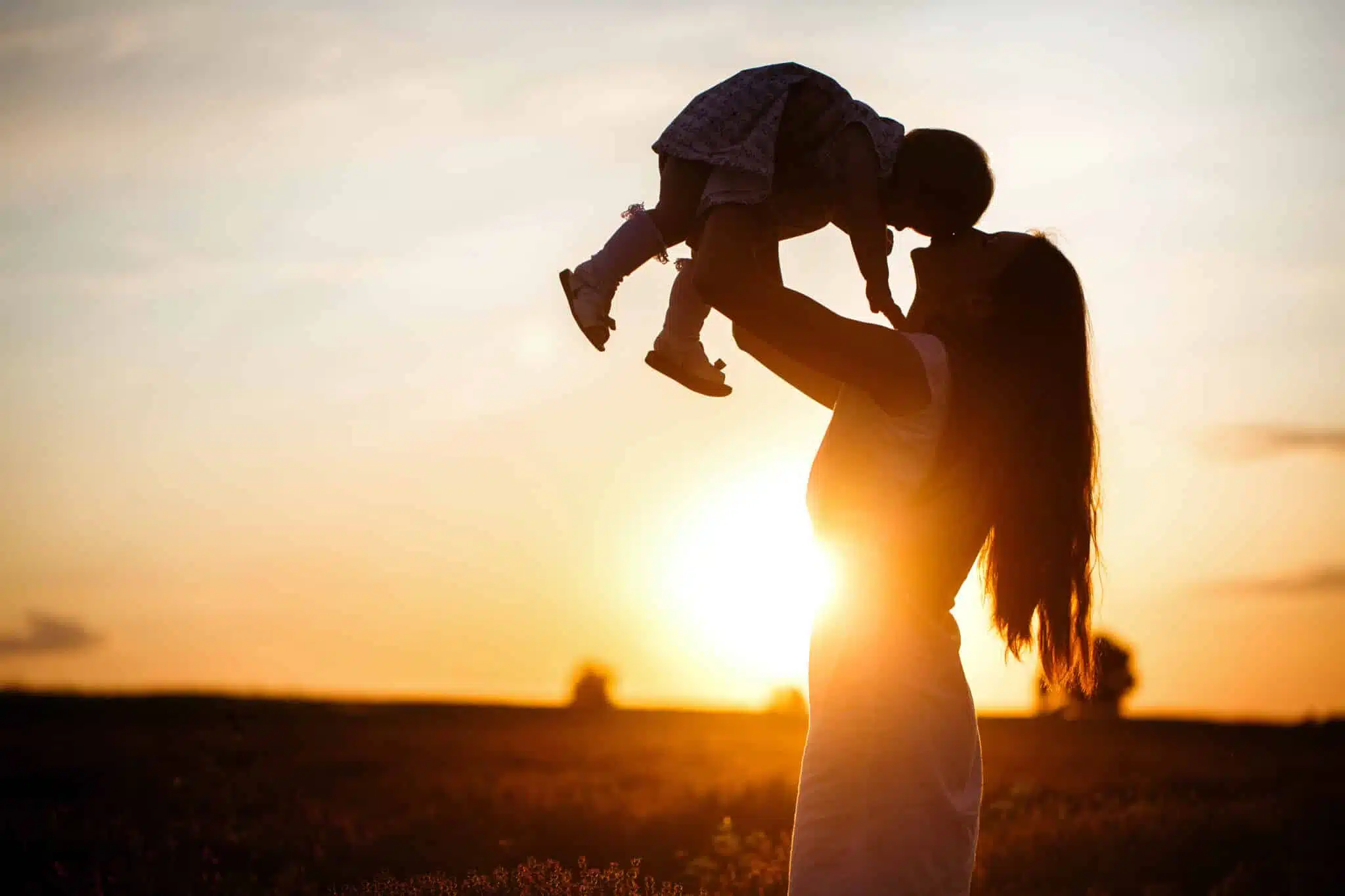 A mother lifts a toddler child in the air above a picturesque sunset sky. A woman and a little girl in a field of lavender flowers
