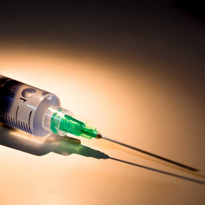 Disposable plastic syringe with needle and blue liquid inside laying on a white surface and illuminated by a selective yellow and white light