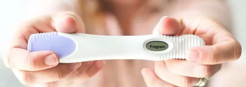 woman in a pink shirt stretching out her arms and holding a positive pregnancy test