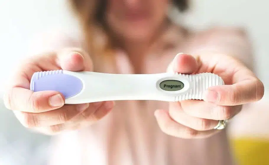 woman in a pink shirt stretching out her arms and holding a positive pregnancy test