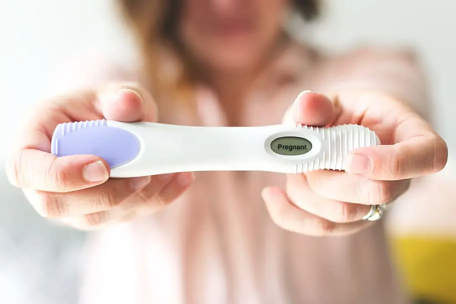 woman in a pink shirt stretching out her arms and holding a positive pregnancy test