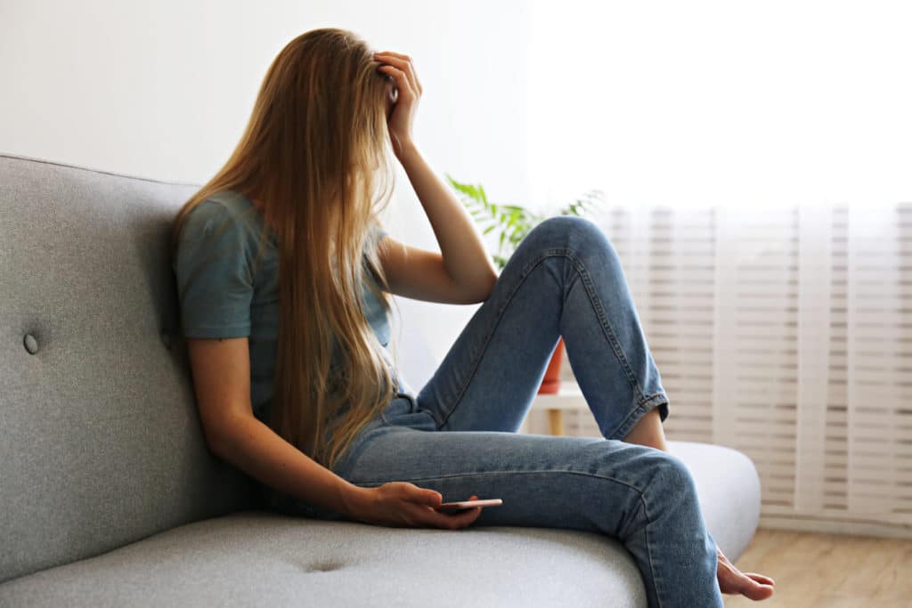 Portrait of depressed young woman hiding her face sitting on grey textile couch holding the phone. Sad female in her room. Female porn addiction, women addicted to porn.