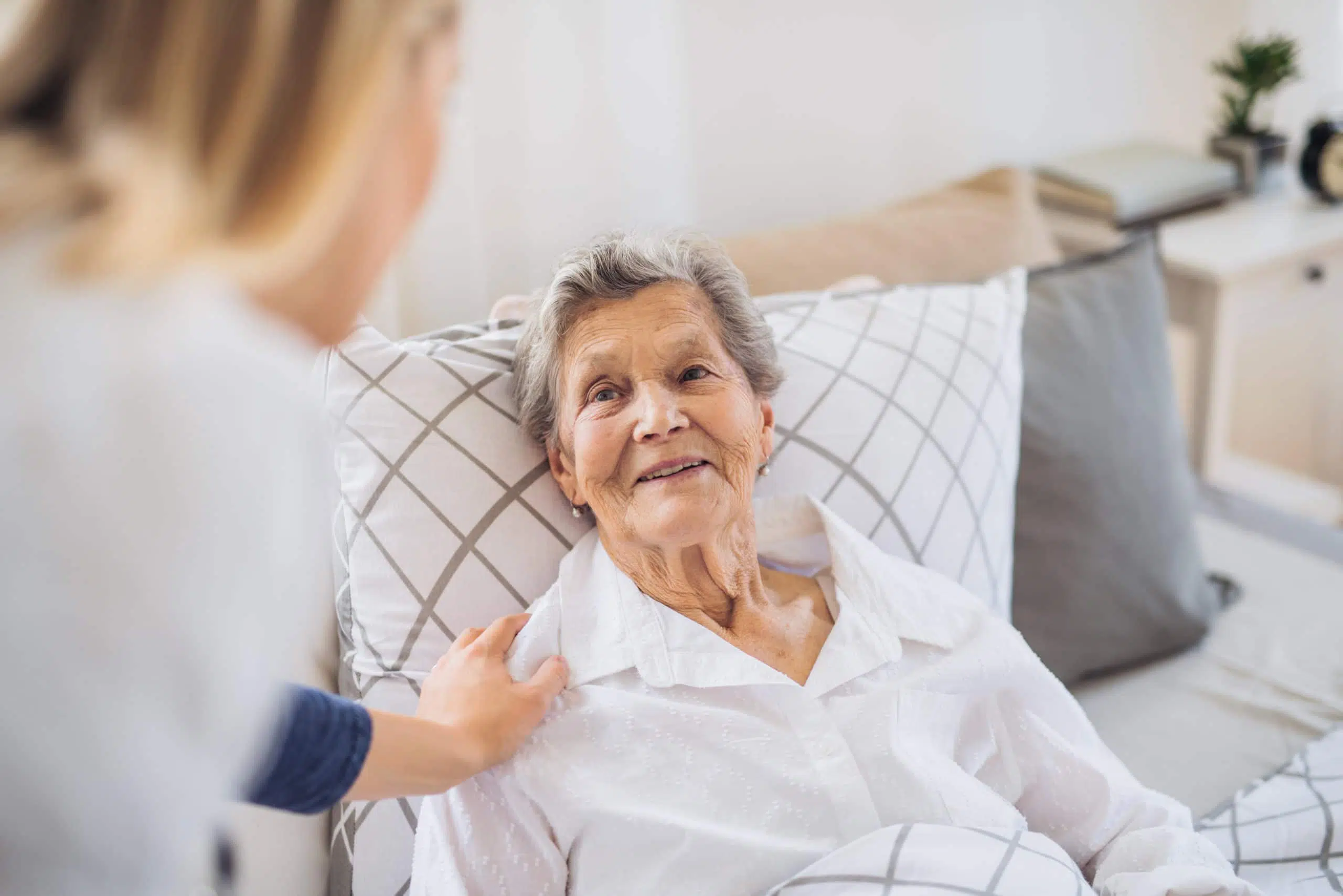 A young health visitor talking to a happy sick senior woman lying in bed at home.