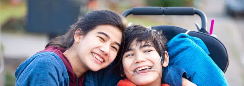 Biracial big sister lovingly hugging disabled little brother in wheelchair outdoors, smiling