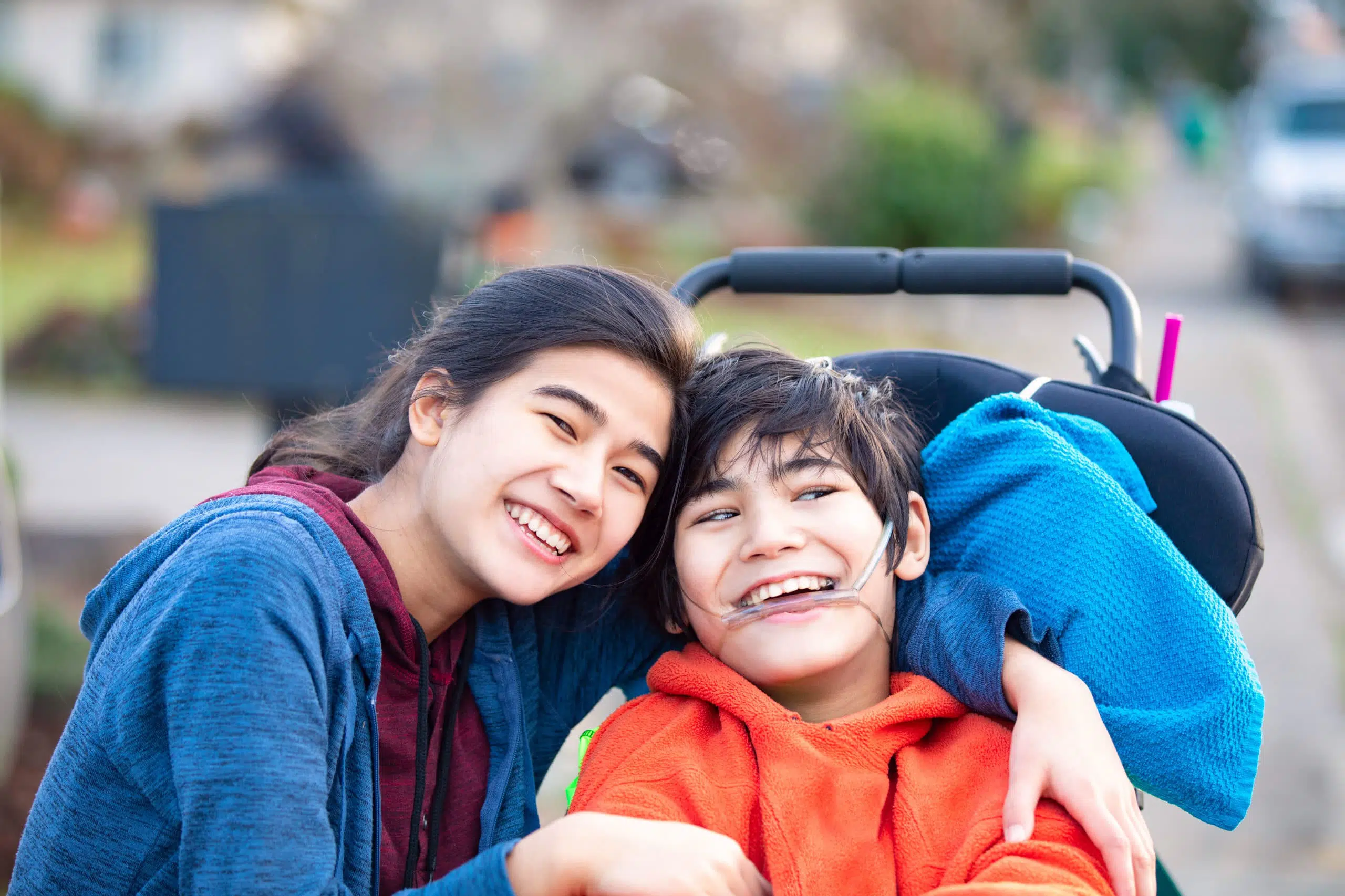 Biracial big sister lovingly hugging disabled little brother in wheelchair outdoors, smiling