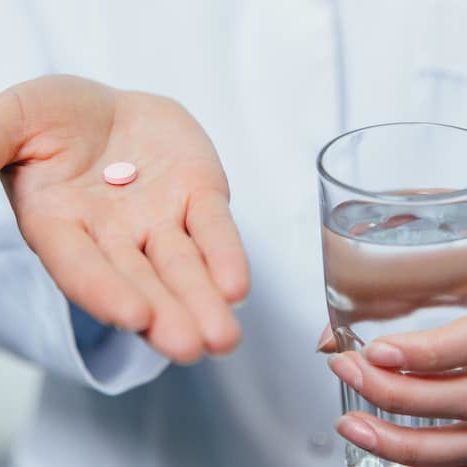 A young girl doctor takes vitamins. At the same time, she stands on a gray background dressed in a white robe. Holds a glass of water and a pill in his hands. On a gray background.