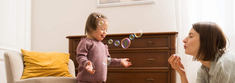 Full length view of the little girl with down syndrome feeling fun while her young mother blowing soap bubbles at home at the morning. Stock photo